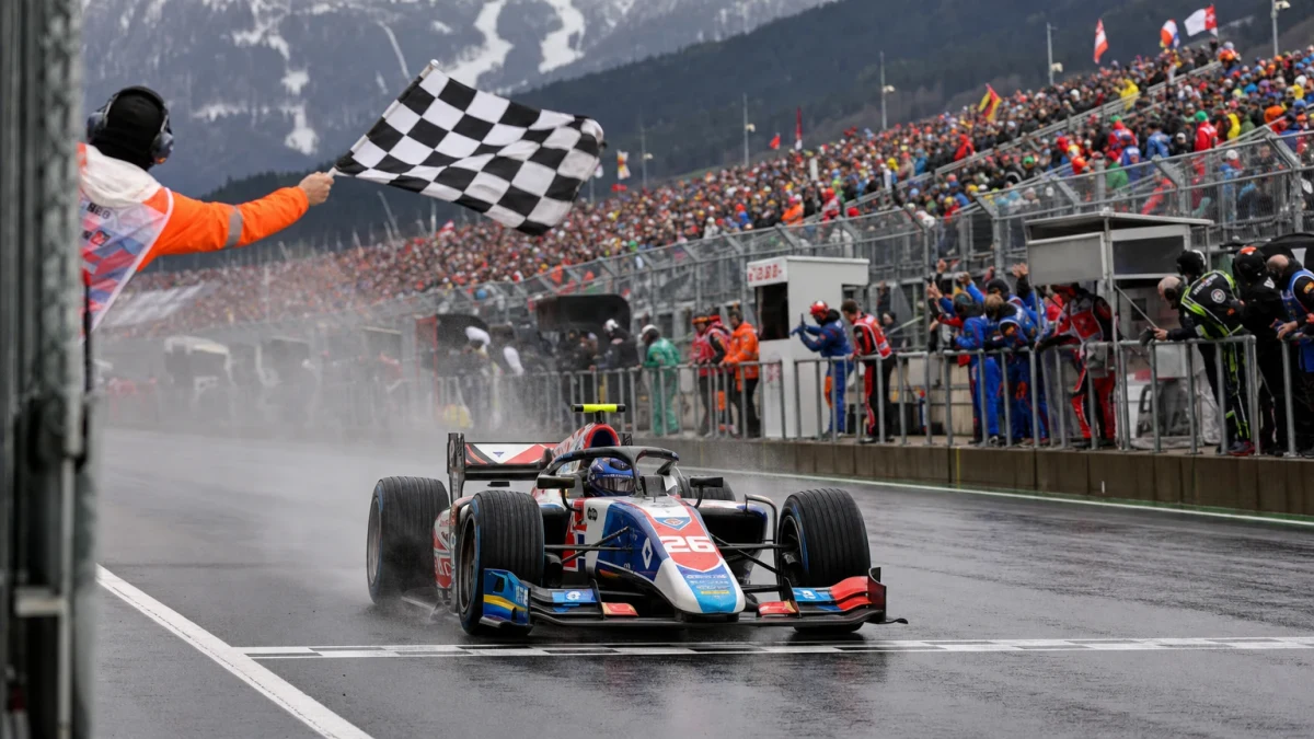 Silverfärgad formel 1-bil leder loppet över mållinjen på en bergig österrikisk racerbana i solljus.